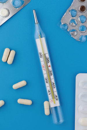 Various pills in packaging, capsules and a medical thermometer on a blue background. Top view,...