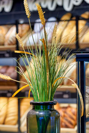 Spikelets of ripe wheat in a glass jar on a blurred background of a showcase with bread in a bakery. Vertical frameの写真素材