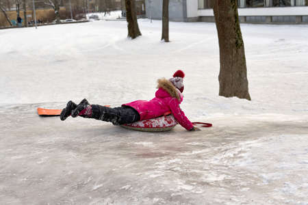 A girl in a red jacket rides on an ice slide on an inflatable sled.の写真素材