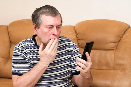 A man looks emotionally into a smartphone while sitting on the couch. Emotions from the newsの写真素材