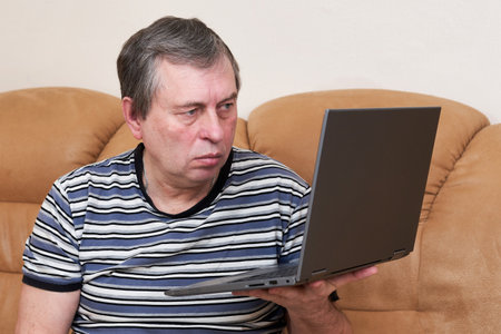 A man with a surprised face holds a laptop in his hands while sitting on couchの写真素材