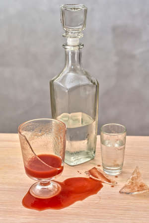 A broken glass, red tomato juice spilled on the table next to a bottle of vodkaの写真素材