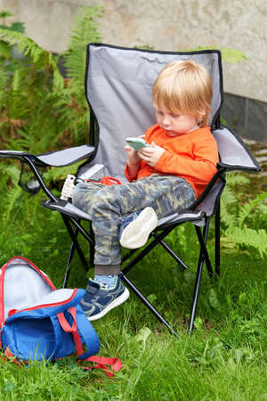A child sits in a tourist chair at a picnic and plays on the phone. Little boy with smartphoneの写真素材