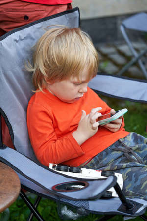 A child sits in a tourist chair at a picnic and plays on the phone. Little boy with smartphoneの写真素材