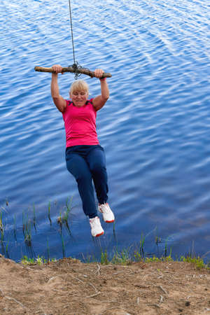 Middle aged woman in sportswear hanging on a stick with a rope above the waterの写真素材