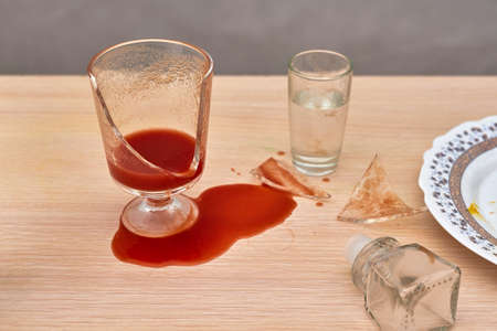 A broken glass, red tomato juice spilled on the table next to a bottle of vodkaの写真素材