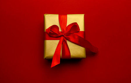 Holiday gift. Box in a gold gift wrapping with a red bow, on a red background. Merry Christmas, Happy New Year and Valentines Day greetings. Postcard to loved ones. High quality photoの写真素材