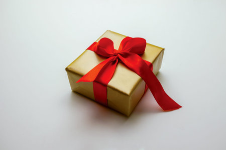 A holiday gift. Box in a gold gift wrapping with a red bow, on a white background. Merry Christmas, Happy New Year and Valentines Day greetings. Postcard. High quality photoの写真素材