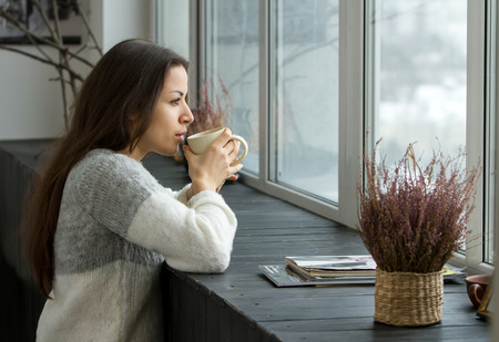 A girl in a bright sweater holding a cup near the lips and looking into the distance through the window, leaning on the wooden sill.の写真素材