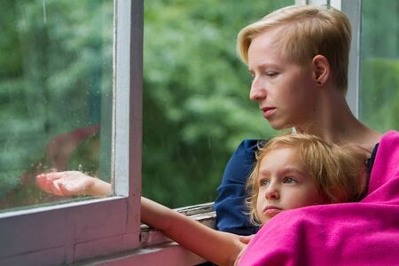 Mother and daughter are sitting by the open window during the rain, the mother holds her daughter in a blanket on herself at that moment when the daughter put her hand out the window and catches raindrops.の写真素材