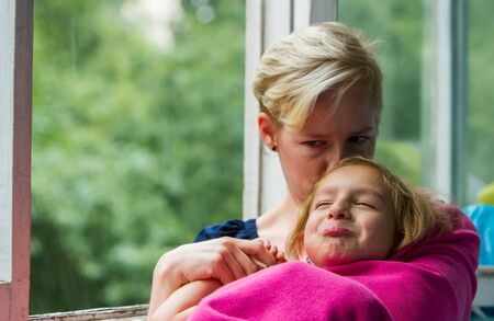 Mother and daughter are sitting near the open window during the rain, the mother holds her daughter in a blanket on herself at the moment when the child is emotional.の写真素材