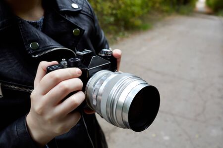 a woman in a leather jacket that holds a film retro camera in her hands, in the background a blurred asphalt alley and green treesの写真素材