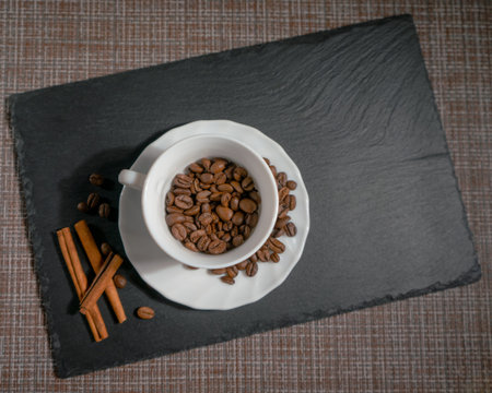 Coffee roasted beans with cinnamon sticks in white ceramic mug brown background