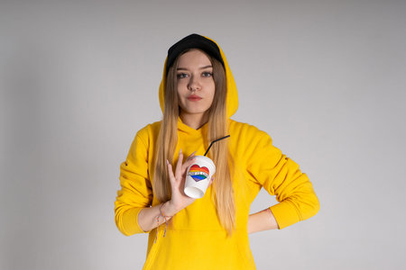 A woman in a yellow hoodie, a black cap with a pierced nose, holds a glass with the inscription love is love and a rainbow LGBTQの写真素材