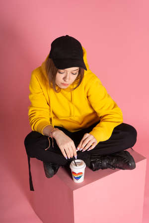 Young LGBTQ sad woman wearing yellow hoodie jacket holding a cup with rainbow heart, on pink backgroundの写真素材