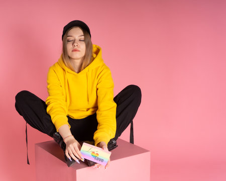 Dreamy girl wearing yellow hoodie jacket and ball cap holding a LGBTQ rainbow love card looking sad or melancholy on pink background.の写真素材