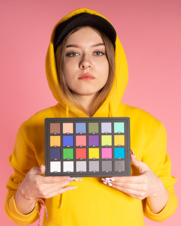 sad girl in a yellow hoodie and a black cap is in the studio holding a color checker in her hands, the background behind her is pinkの写真素材