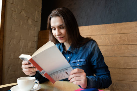 a cheerful girl in a blue denim shirt sits in a cafe and reads a book