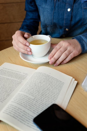 hands holding a white glass with green tea, in the foreground lies a book outside the sharpness...