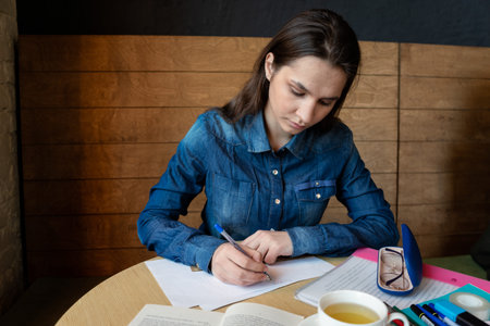 serious girl student writes with a black pen, on the table are glasses, flavors, a book and there is a glass of green teaの写真素材