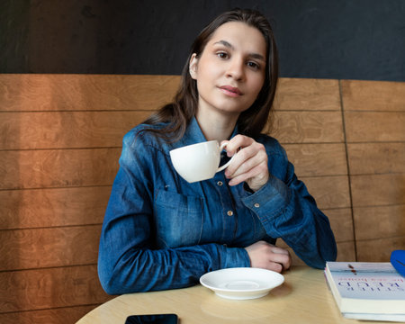 A girl in a blue denim shirt sits in a cafe and looks at a lens with a mug of green tea in her hands