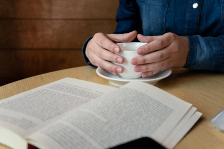 hands holding a white glass with green tea, in the foreground lies a book outside the sharpness...