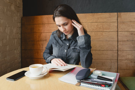 A thoughtful girl in a blue denim shirt sits in a cafe and reads a book, pens and pencils are on the...