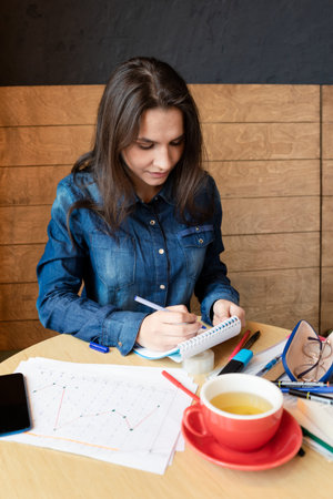 Girl in a blue denim shirt sitting in a cafe makes notes in a notepad with a pen. A smartphone,...