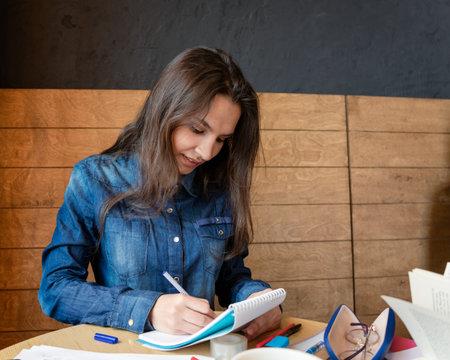 A cheerful girl in a blue denim shirt sitting in a cafe makes notes in a notepad with a pen. Sheets,...