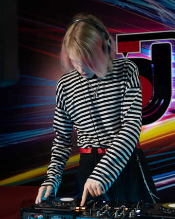 DJ with painted lips, pressing a button on a mixing console, DJ equipment in front of him, wearing a striped hoodie, red belt, is in a nightclubの写真素材