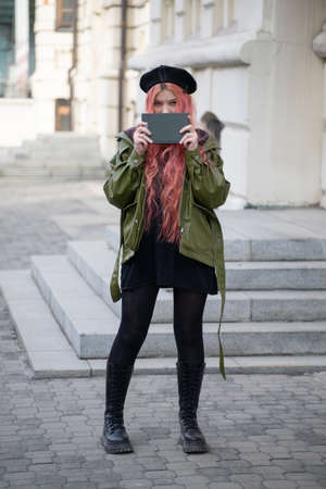 Girl in a black beret, with pink long hair, holding a closed green book near her face, on the street in the city, full growth, she is wearing high black boots, tights, sweatshirt, green jacketの写真素材