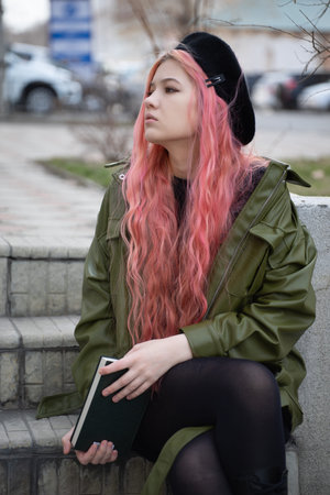 A fashionable student in a black beret with long pink hair sits on the steps and enthusiastically...