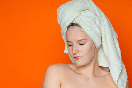 A beautiful sad girl in a cosmetic mask looks down, is on the right side of the frame, has wet hair on which there is a white towel, copy space. Photography in the studio, on an orange backgroundの写真素材