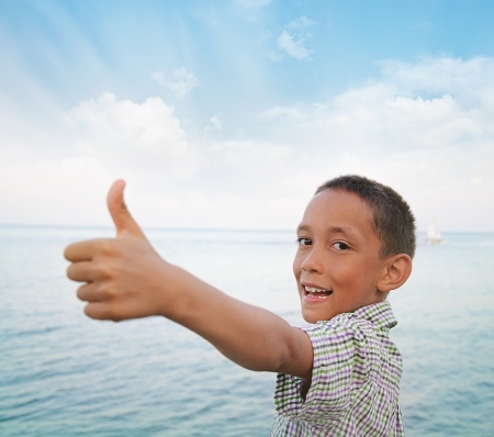 happy sun-tanned boy showing thumbs-up against sea and sky in summerの写真素材