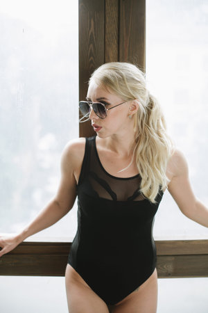 girl in sexy body posing next to a window holding her hands near the sunglasses