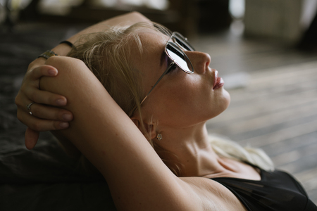 woman with bare feet in black sneakers and body sits near a large bed on the wooden floor, looking at the camera and posing for a photograph with his hands near the face. woman posing beside the bed and holding his hands behind his headの写真素材