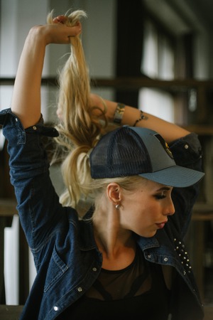 cute long-haired girl in jeans jacket and baseball cap holds for blond hair. Pulls hands behind his tail and looking at the camera.の写真素材