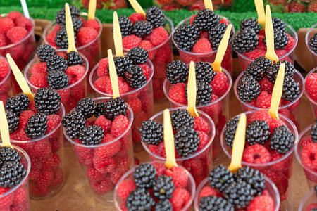 Raspberries and blackberries mix in plastic glasses with spoons on the market counterの写真素材