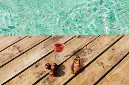 A glass with fruit juice, sun glasses and some plums on wood boards along the edge of a pool with blue mosaic tiles. Summer vibes and leisure minimal composition.の写真素材