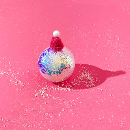 A big shiny Christmas ball with a pink pompom cap on pink background with snow around. Christmas or New Year party invitation creative concept. Surreal festive design.の写真素材
