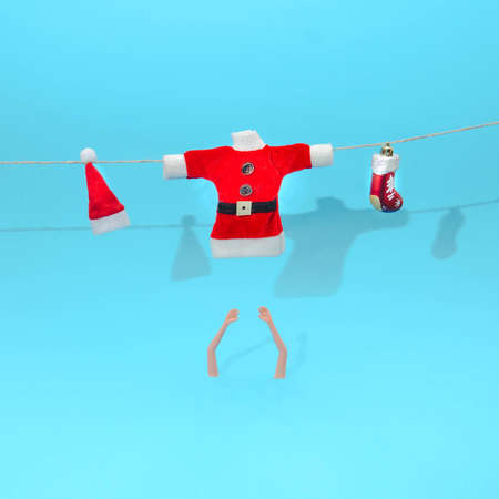 Santa Claus blouse, red hat with white pompom and a red shoe hanging on a rope. Women's arms below. Pastel blue background. Surreal Christmas or New Year's eve party invitation.  Design for card.の写真素材