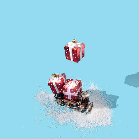 An old red train in the snow transporting two red wrapped gift boxes, a third one is falling from above. Pastel blue background. Retro Christmas or New Year creative composition. Artistic concept for card or banner.の写真素材