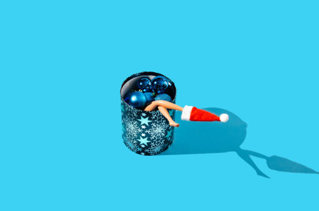 Women's bare legs going out of a round shaped blue Christmas box full of blue baubles, Santa's red hat on one leg. Pastel blue background. Creative concept for Christmas or New Year's party invitation. Festive season minimal advertisement design.の写真素材