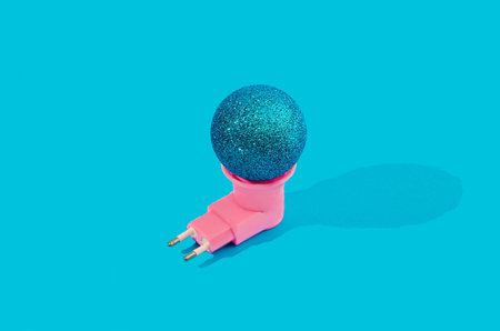 A shiny blue Christmas bauble plugged on a pink power socket. Pastel blue background. Minimal...