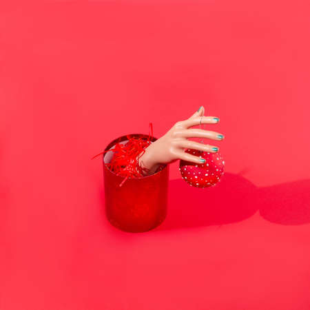 A women's hand with green painted nails going out of a red Christmas box full of tinsels and holding a red bauble. Red background. Creative surreal Christmas or New Year design for card or banner.の写真素材