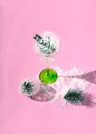 A wine glass with green foot and a Christmas bauble on pink table with snow around, another bauble and a small Christmas tree next to the glass. Romantic concept for festive invitation cardの写真素材