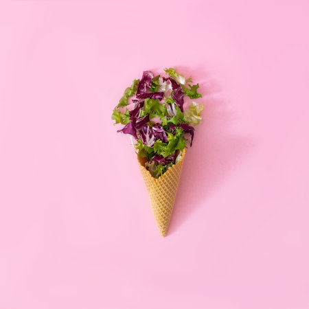 An ice cream cone with fresh green and red salad leafs on pastel pink background. Minimal concept...