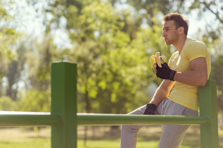 Young sportsman sitting on a bar in an outdoor gym, eating a banana