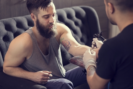 Male tattoo artist holding a tattoo gun, showing a process of making tattoos on a male tattooed model's arm.の写真素材