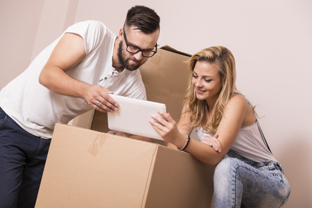 Young couple in love moving in a new apartment, standing next to cardboard boxes, holding tablet computer and surfing the web while planning to redecorate their new homeの写真素材